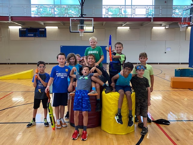 Group of children smiling and posing with foam dart blasters on padded blocks in a gymnasium.